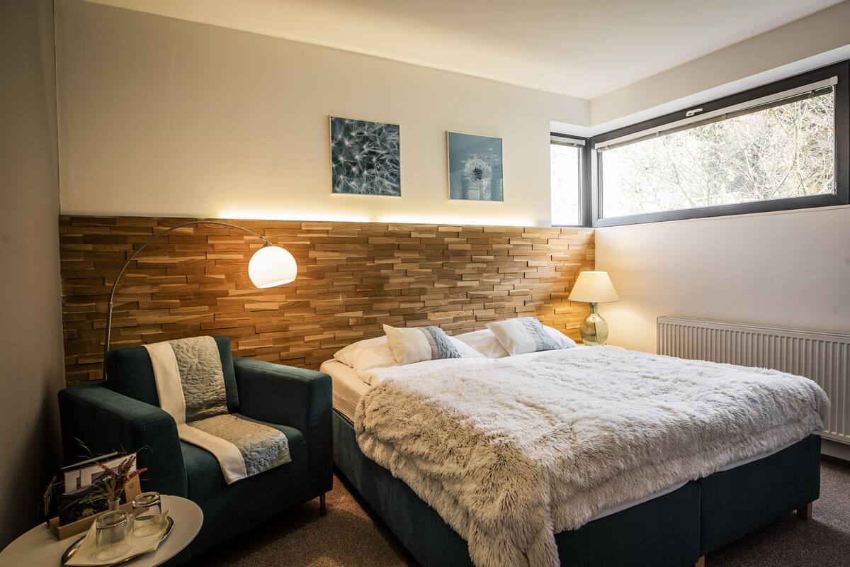 Ein Schlafzimmer mit einem hölzernen Kopfteil, einem Bett mit flauschiger weißer Decke, einem grünen Sessel, einem Nachttisch mit Lampe und zwei gerahmten Bildern an der Wand.