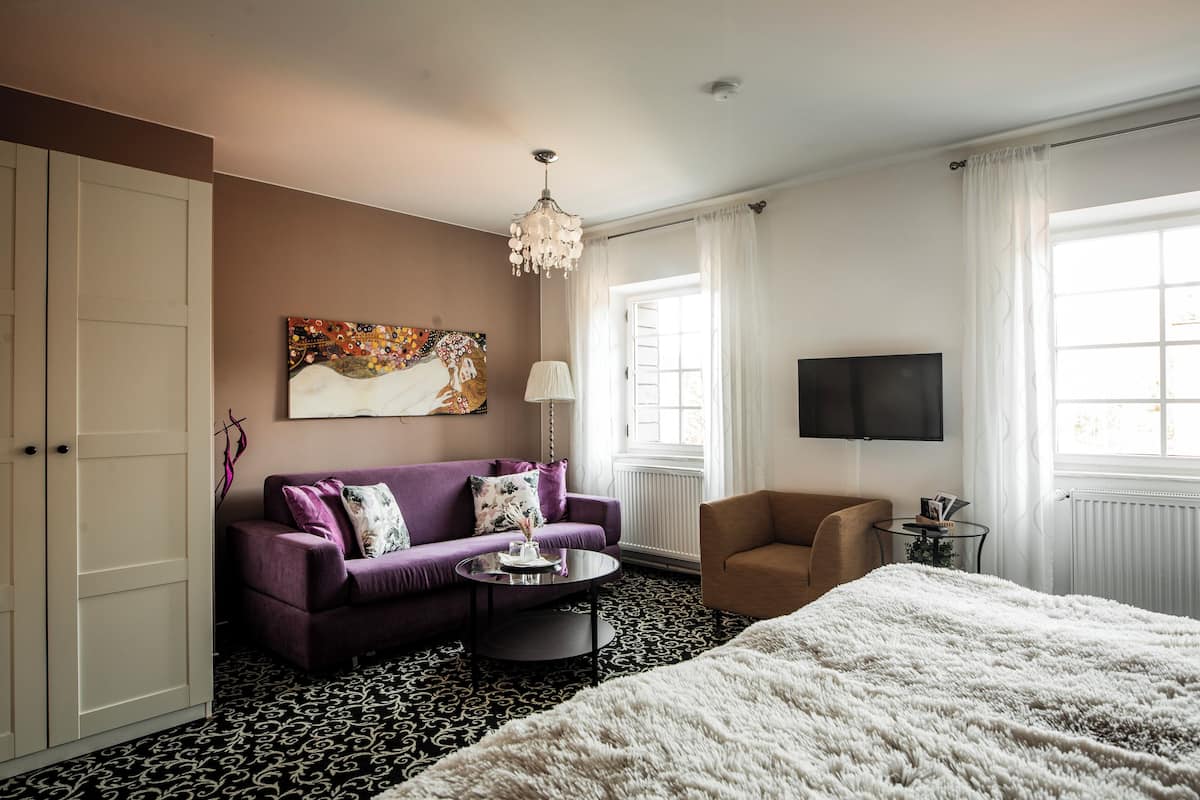 Ein Hotelzimmer mit einem Bett, einem violetten Sofa, einem runden Couchtisch, einem braunen Sessel, einem Fernseher und einem großen Fenster mit Vorhängen.