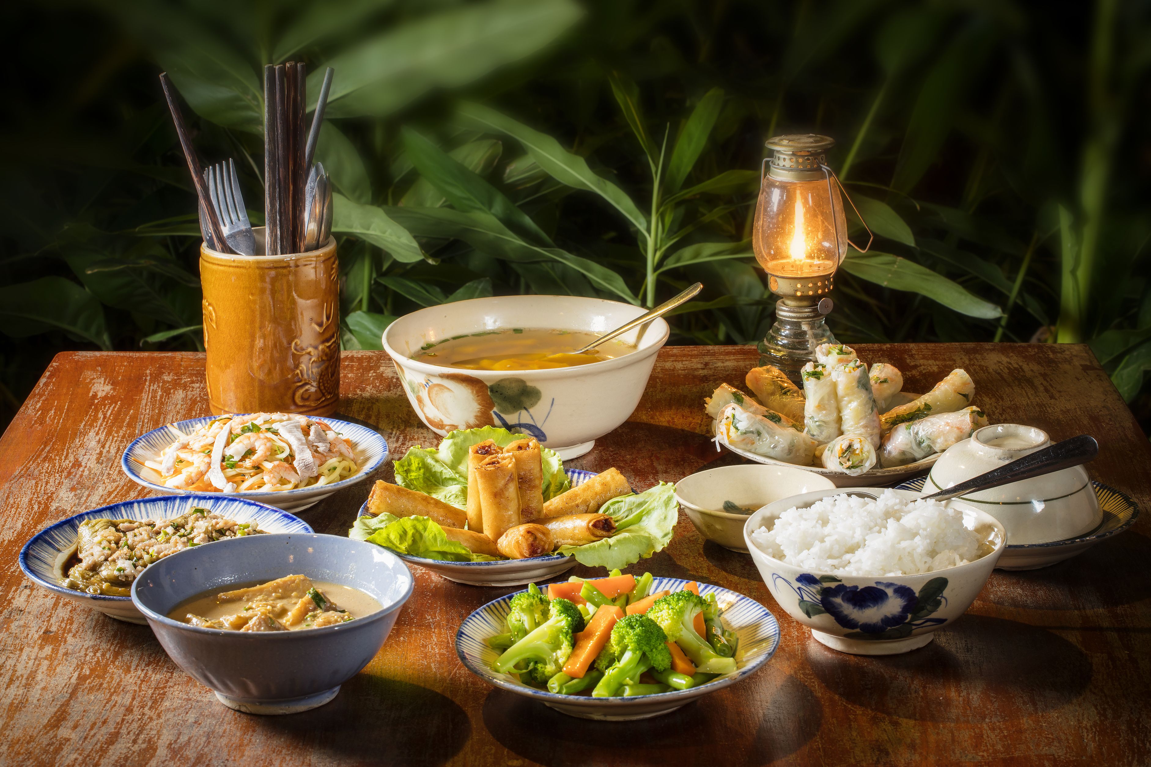 Une table dressée avec divers plats, notamment du riz, des légumes et des rouleaux de printemps, le tout mis en valeur par un décor de feuillage verdoyant.