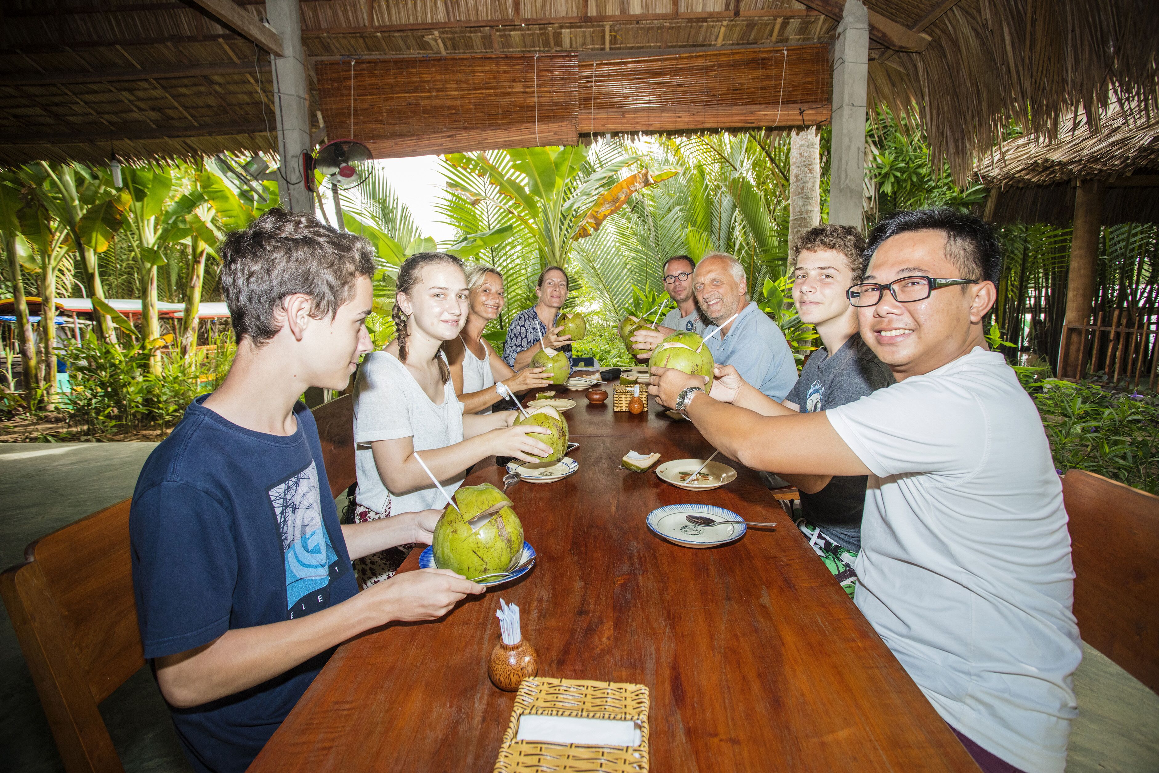 A group of people sitting around a wooden table outdoors, holding coconuts.
