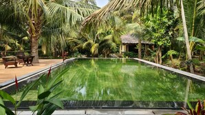 A rectangular swimming pool surrounded by lush greenery and palm trees, with a thatched-roof structure in the background. - Mekong Home (Phuoc Long)