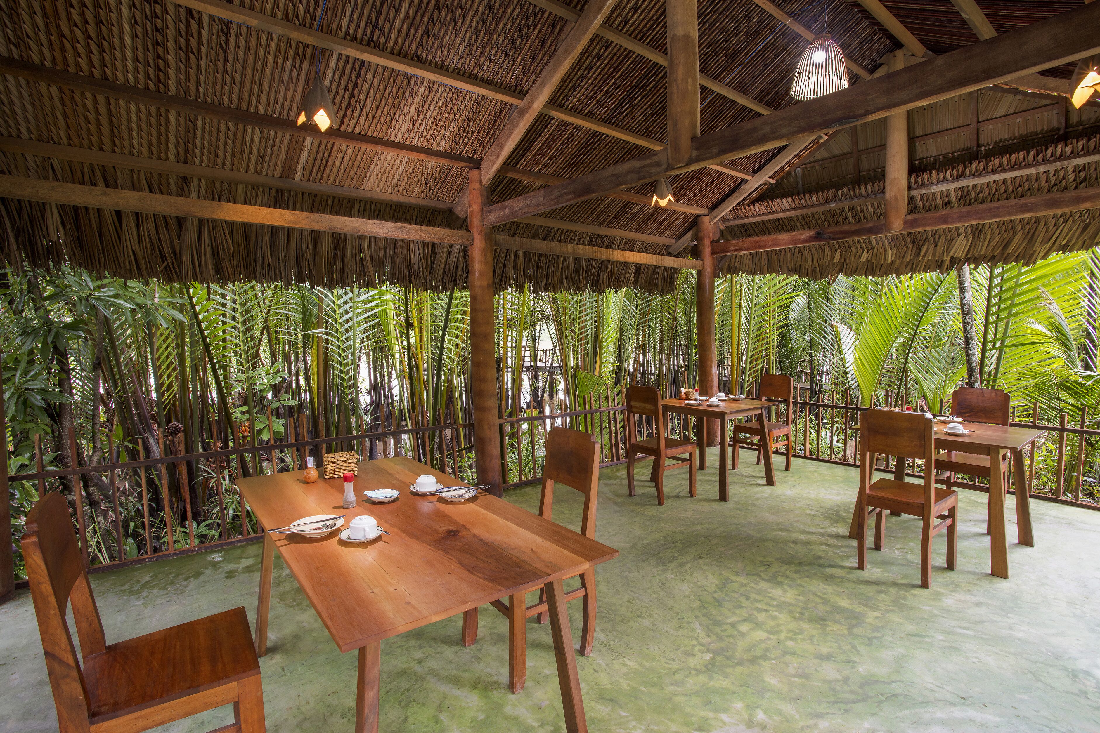 Un espace de restauration en plein air avec des tables et des chaises en bois, entouré d’une végétation luxuriante et surmonté d’une structure à toit de chaume.