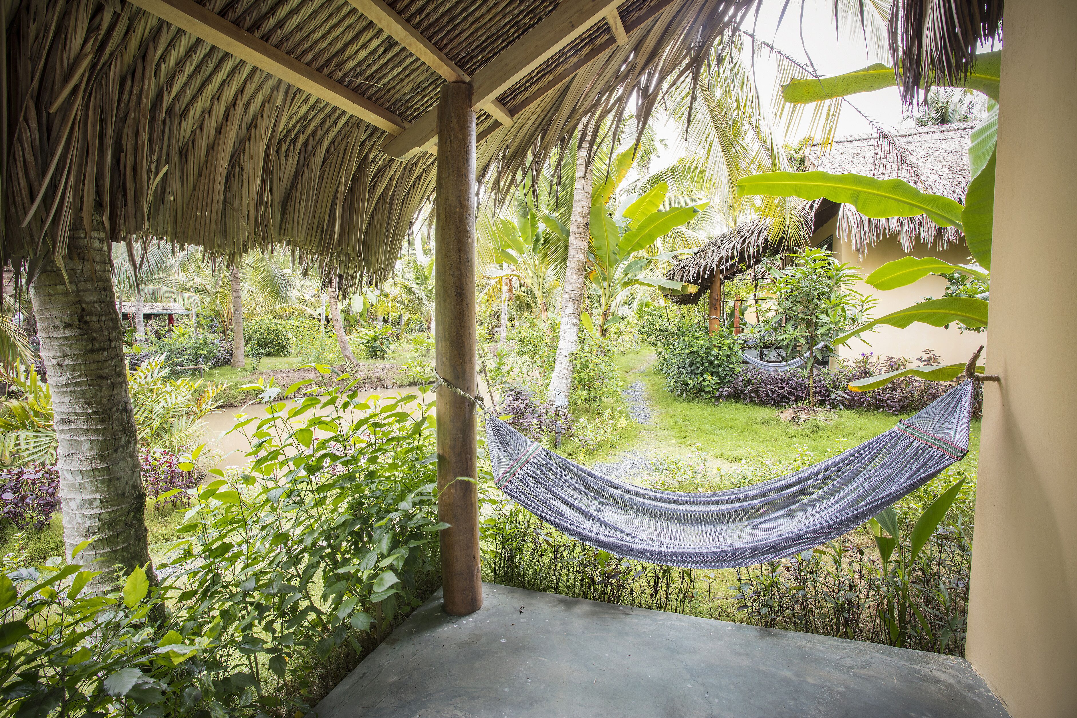 A hammock under a thatched roof structure, surrounded by lush greenery and a garden.