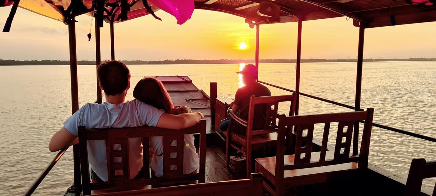 A boat with wooden benches and a canopy, passengers facing the sunset over water.