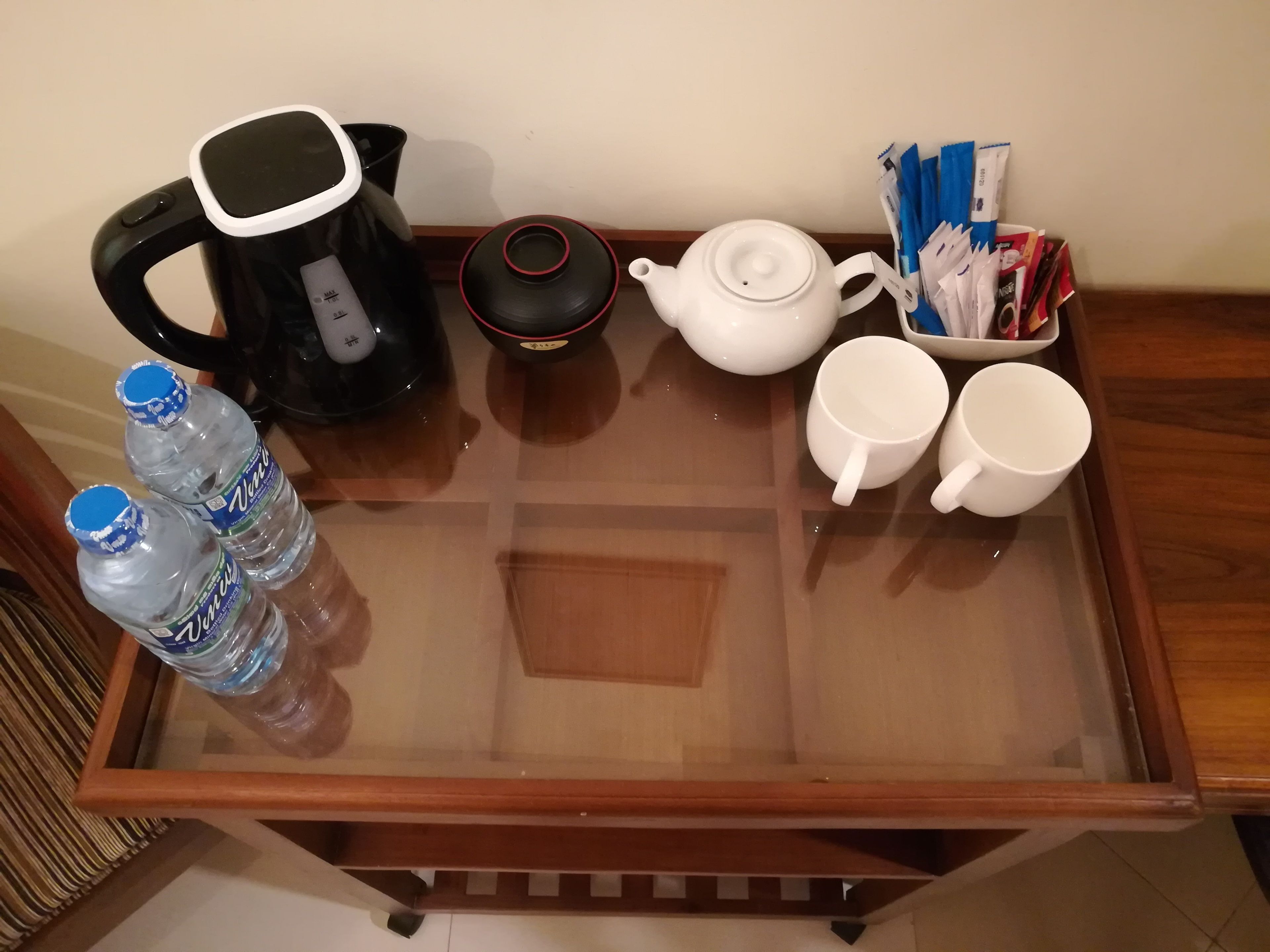 A glass-top table with a black electric kettle, a white teapot, two white cups, and two bottles of water.