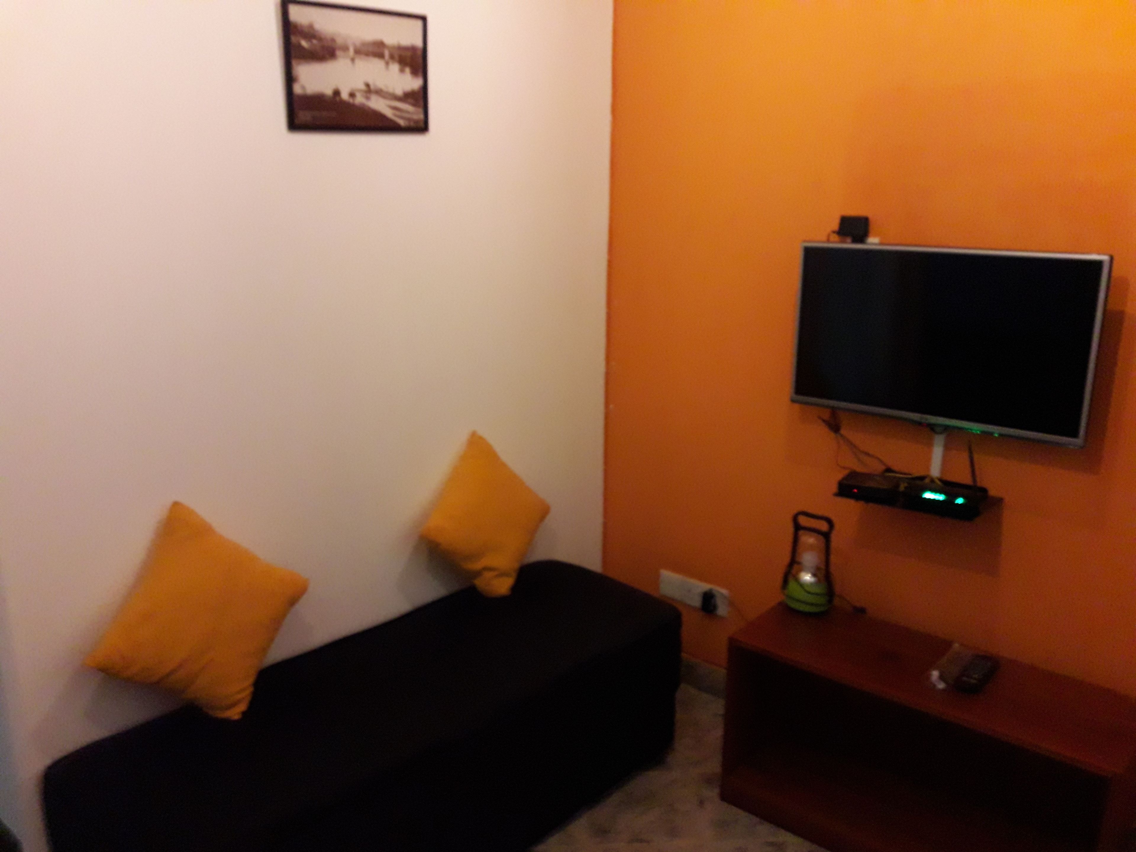 A living room with a black sofa, two yellow cushions, a flat-screen TV mounted on an orange wall, and a small wooden side table with a remote control.