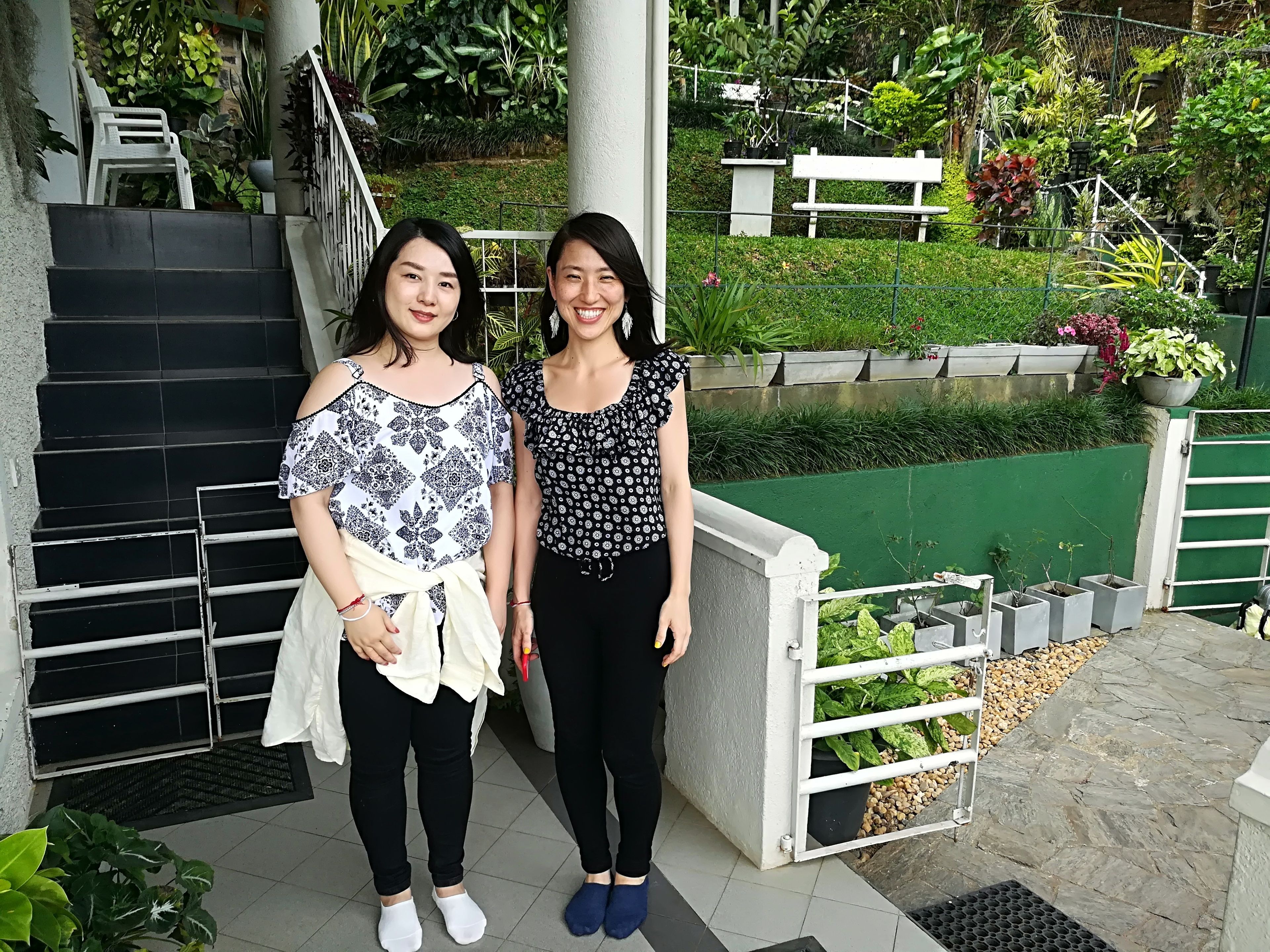 Two individuals standing on a staircase with a green garden and a white bench in the background.