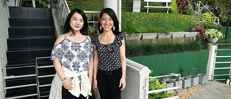 Two individuals standing on a staircase with a green garden and a white bench in the background.