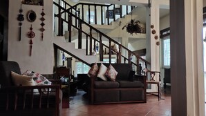 Amplia sala de estar con un sofá marrón oscuro, una mesa de centro de madera y una silla. Una escalera con barandas de madera conduce a un nivel superior.