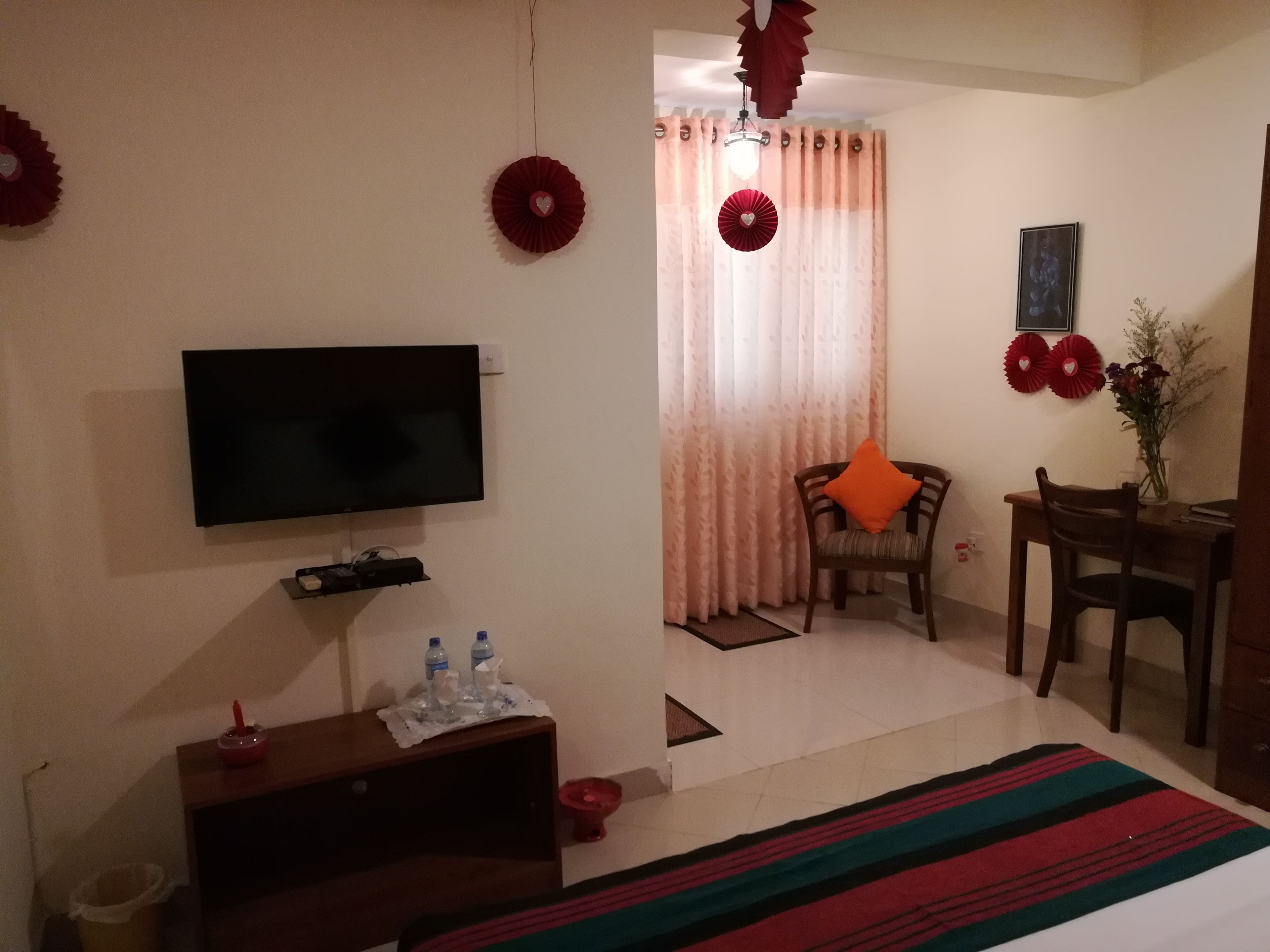 Ein Hotelzimmer mit Flachbildfernseher, einem Holznachttisch mit Vase und rotem Kissen, einem Essbereich mit Tisch und Stühlen sowie einem gemusterten Teppich.
