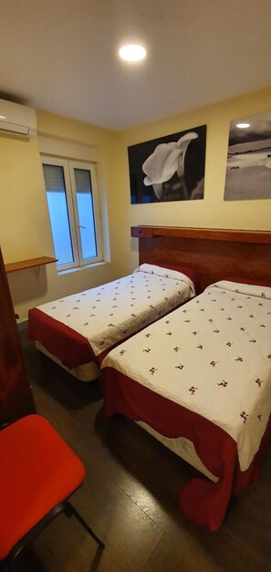 A room with two beds, a red chair, a window, and a wall-mounted air conditioner. - Hostal Moncloa (Madrid)