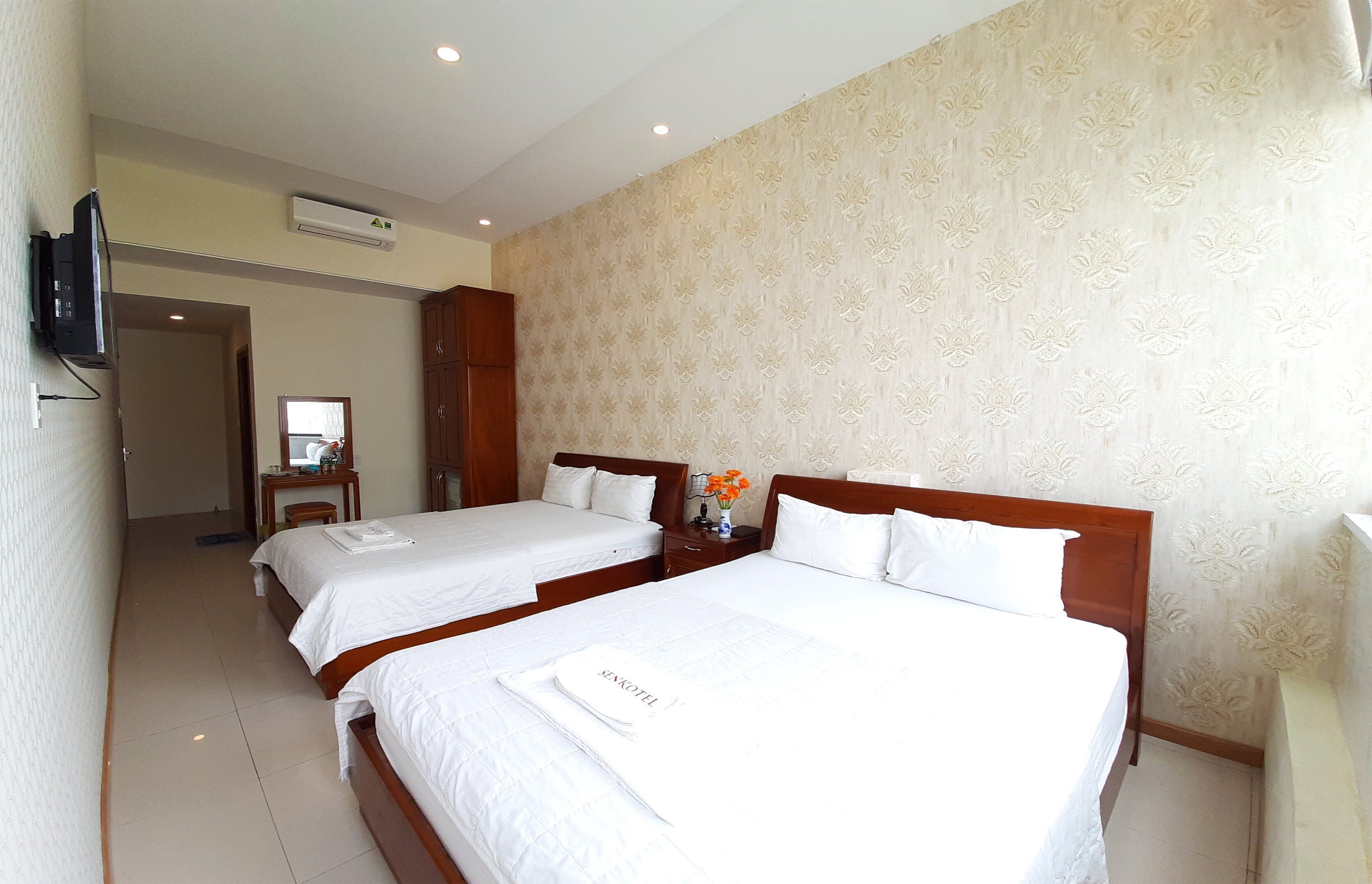 Habitación de hotel con dos camas, televisor, espejo y una mesita con un jarrón de flores.
