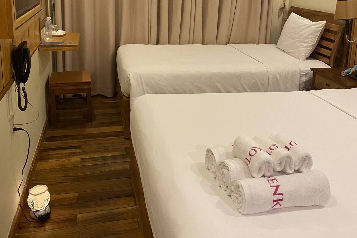 A hotel room with two beds, a wooden floor, a small table with a phone, and a window with curtains.