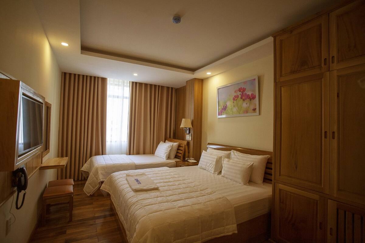 A hotel room with two beds, a wooden wardrobe, a desk with a lamp, and a painting on the wall.
