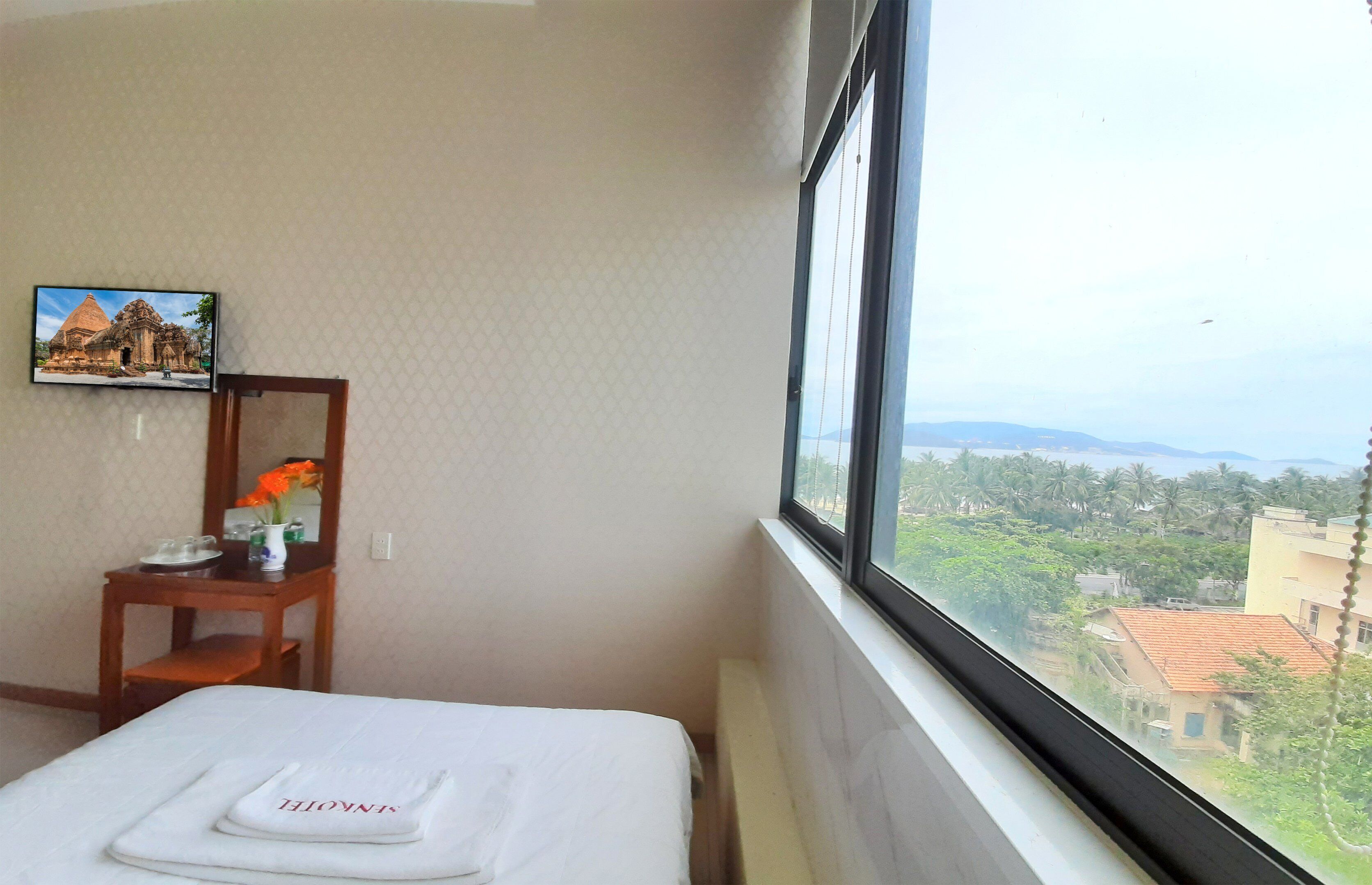A hotel room with a bed, a small table with a vase of flowers, a framed picture on the wall, and a window with a view of buildings and greenery.