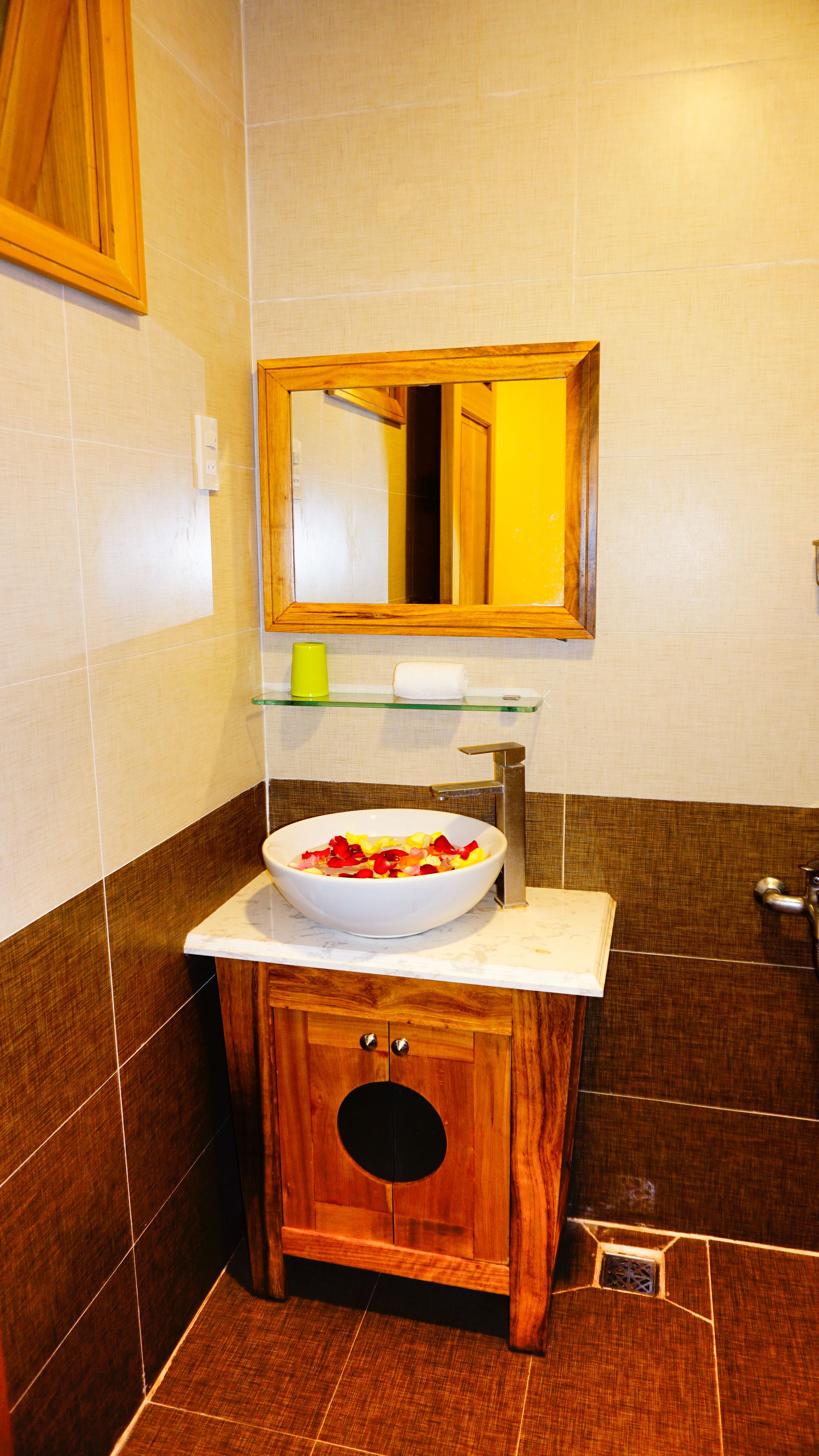 Un baño con tocador de madera, un lavabo blanco con cuenco, un espejo sobre el lavabo y una repisa de cristal con una taza amarilla y un rollo de papel higiénico.