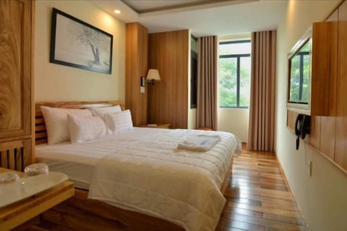 A hotel room with a bed, bedside table, lamp, window with curtains, and a painting on the wall.