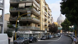 A multi-story building with balconies, a street view, parked cars, and a dome structure in the background. - Beside the Vatican (Rome)