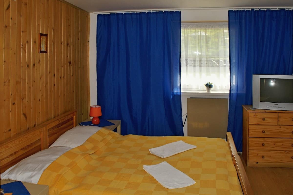 A hotel room with a wooden wardrobe, a bed with a yellow checkered bedspread, a television on a wooden dresser, and blue curtains.