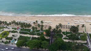 Eine Vogelperspektive zeigt einen Strand mit mehreren Reihen von Sonnenschirmen, Palmen und einem Parkplatz mit Motorrädern.