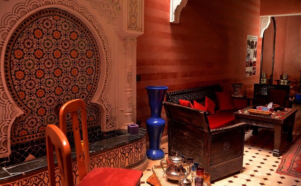 Una habitación marroquí tradicional con intrincados trabajos en azulejos, un sofá de madera con cojines rojos, un jarrón azul y una pequeña mesa con una tetera y vasos.