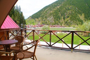 Un balcón con sillas de mimbre y una mesa, con vista a un paisaje montañoso y edificios a lo lejos.