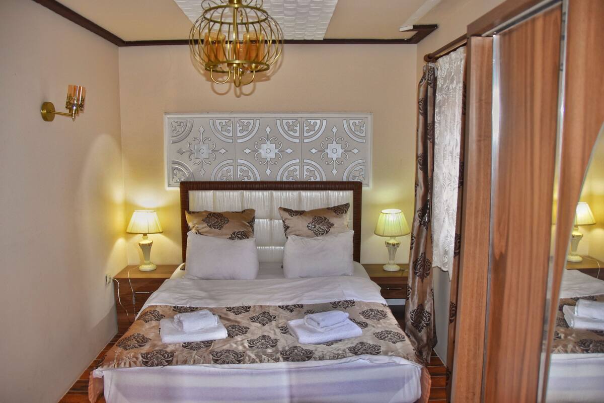 A neatly made bed with a patterned bedspread, two bedside lamps, a decorative wall panel, and a chandelier.
