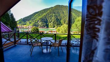 A balcony with wicker chairs and a table, overlooking a mountainous landscape with a lake and buildings.