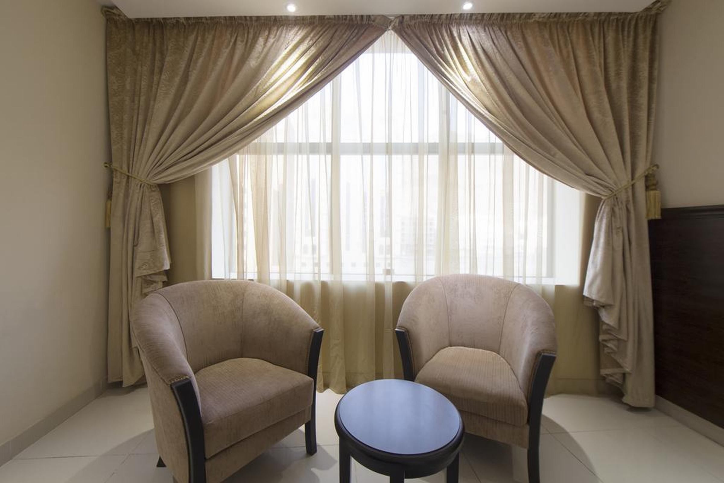 A room with two armchairs and a small table, a large window with sheer curtains, and a dark-colored television on the right.