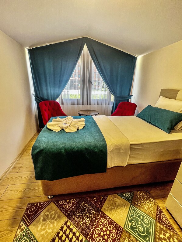 Habitación de esquina con una cama, dos sillas rojas, una mesita redonda y vistas por una ventana con cortinas azules.