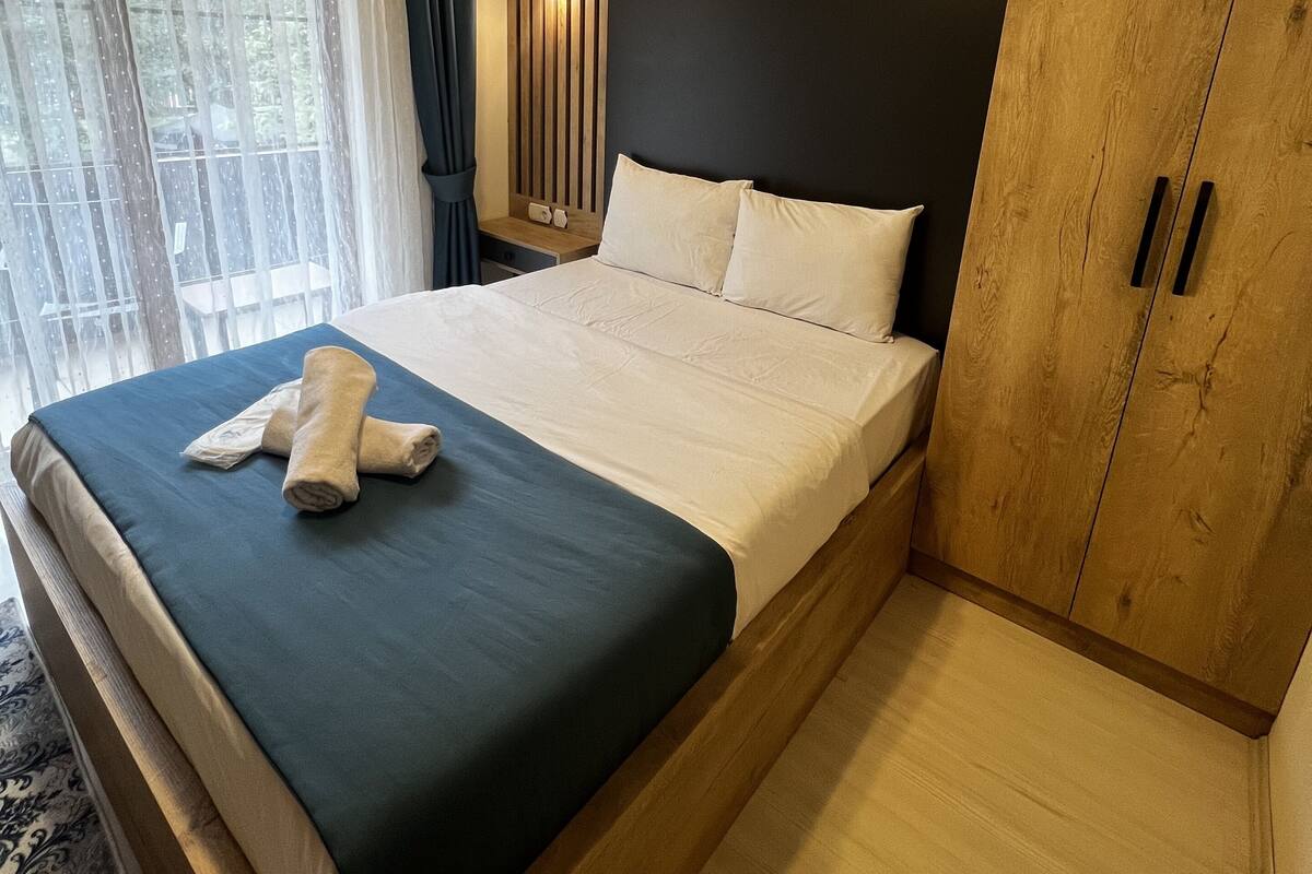A hotel room with a large bed, a wooden wardrobe, a bedside table with a mirror, and a view of greenery through the window.