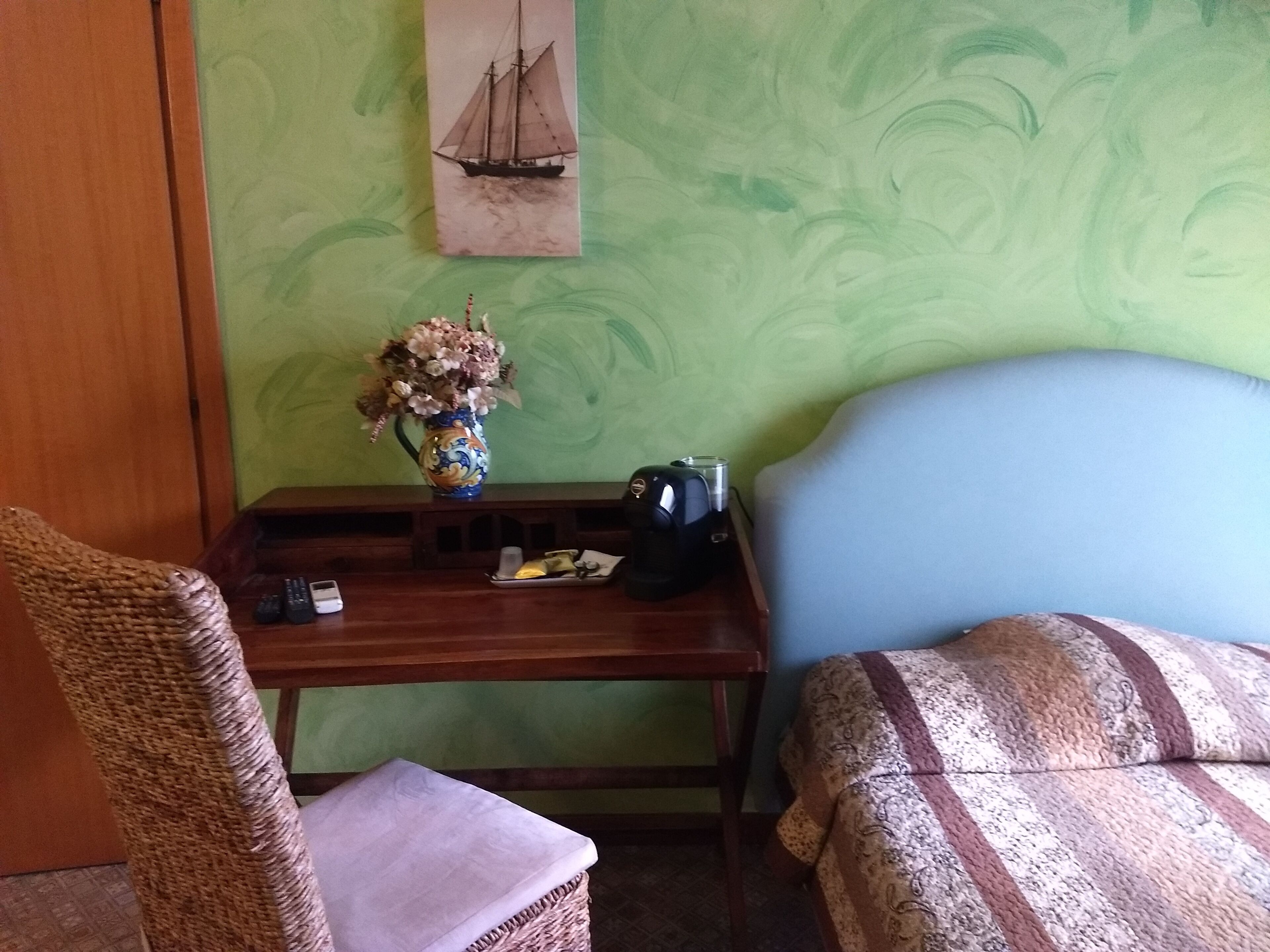 Quarto com escrivaninha de madeira, um vaso de flores, pintura de um navio e cama com roupa de cama estampada.
