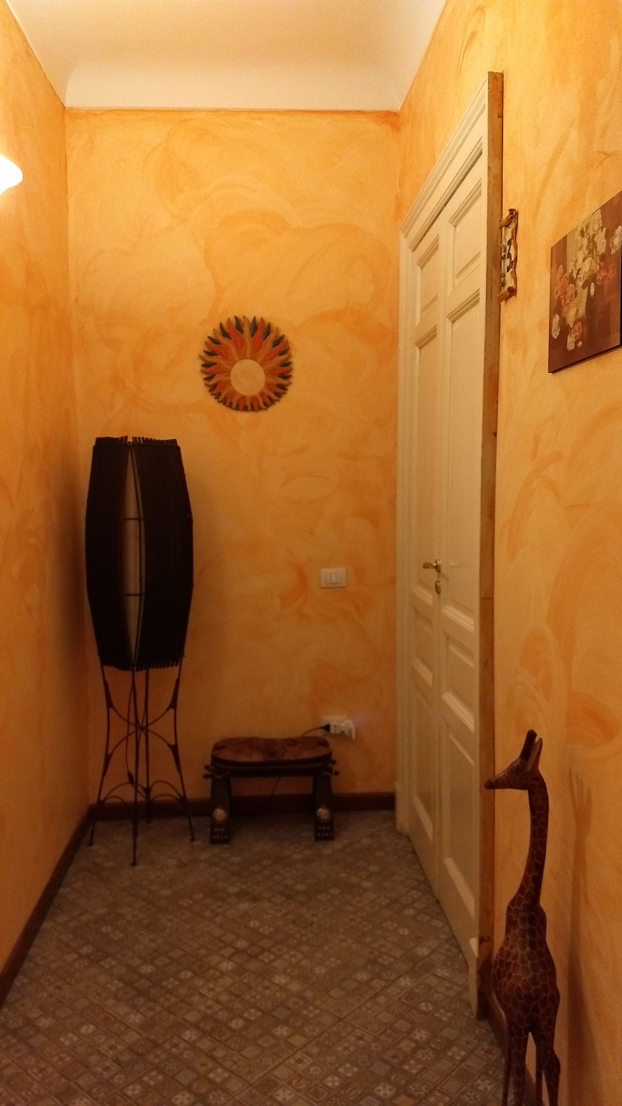 A narrow hallway with a patterned floor, a small table with a stool, a decorative sun-like object on the wall, and a giraffe figurine.