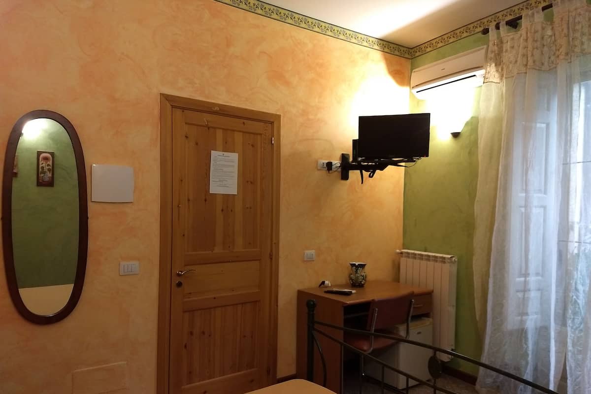 Una habitación con puerta de madera, una pequeña mesa con un jarrón, televisor montado en la pared y ventana con cortina.