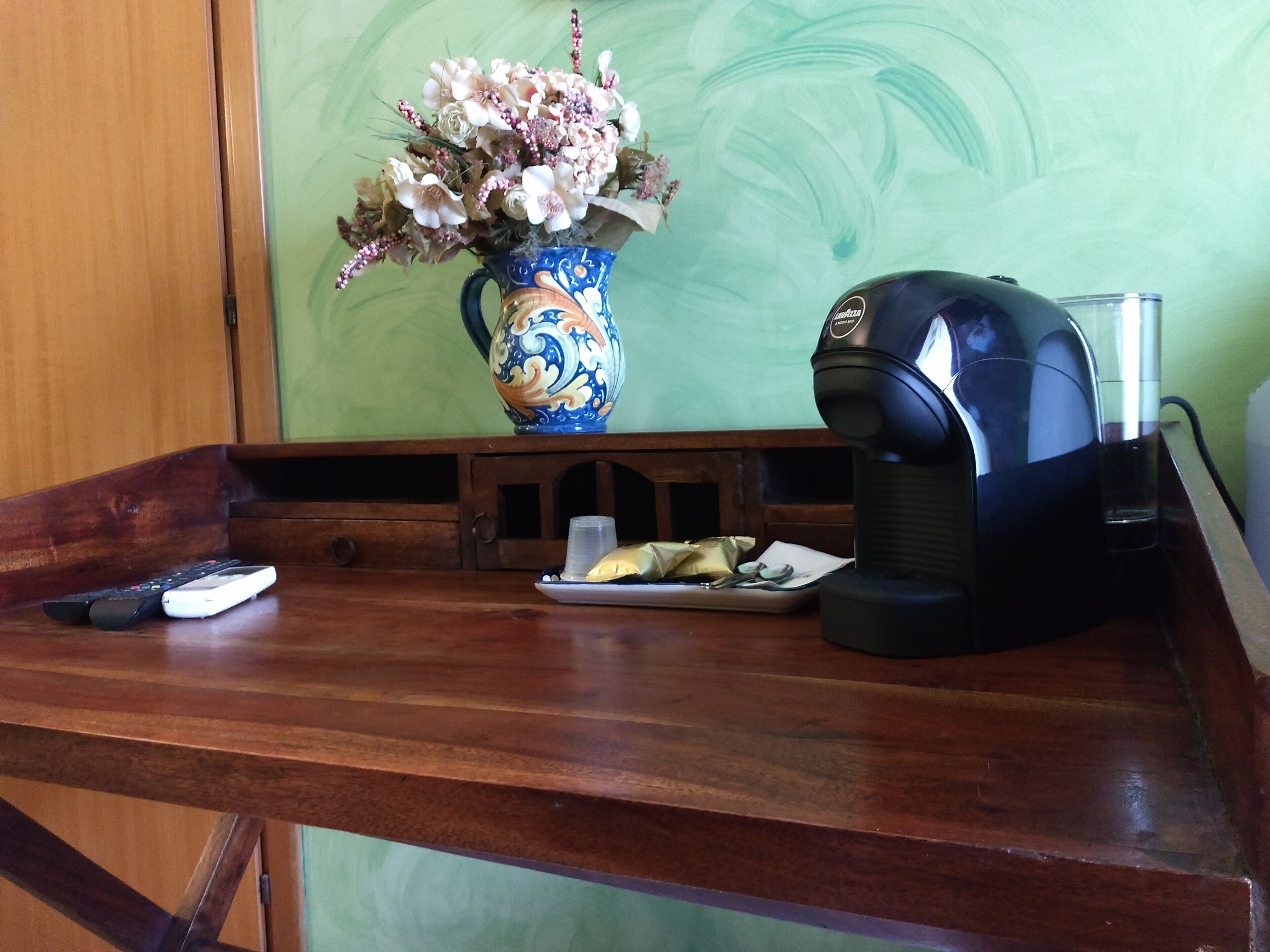 A wooden desk with a vase of flowers, a coffee maker, and a plate with food.
