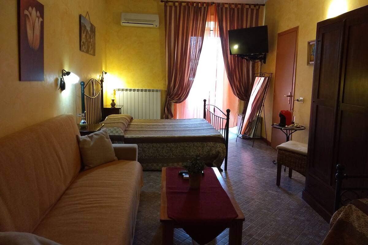 Una habitación con cama, sofá, televisor, mesa y ventana con cortinas.