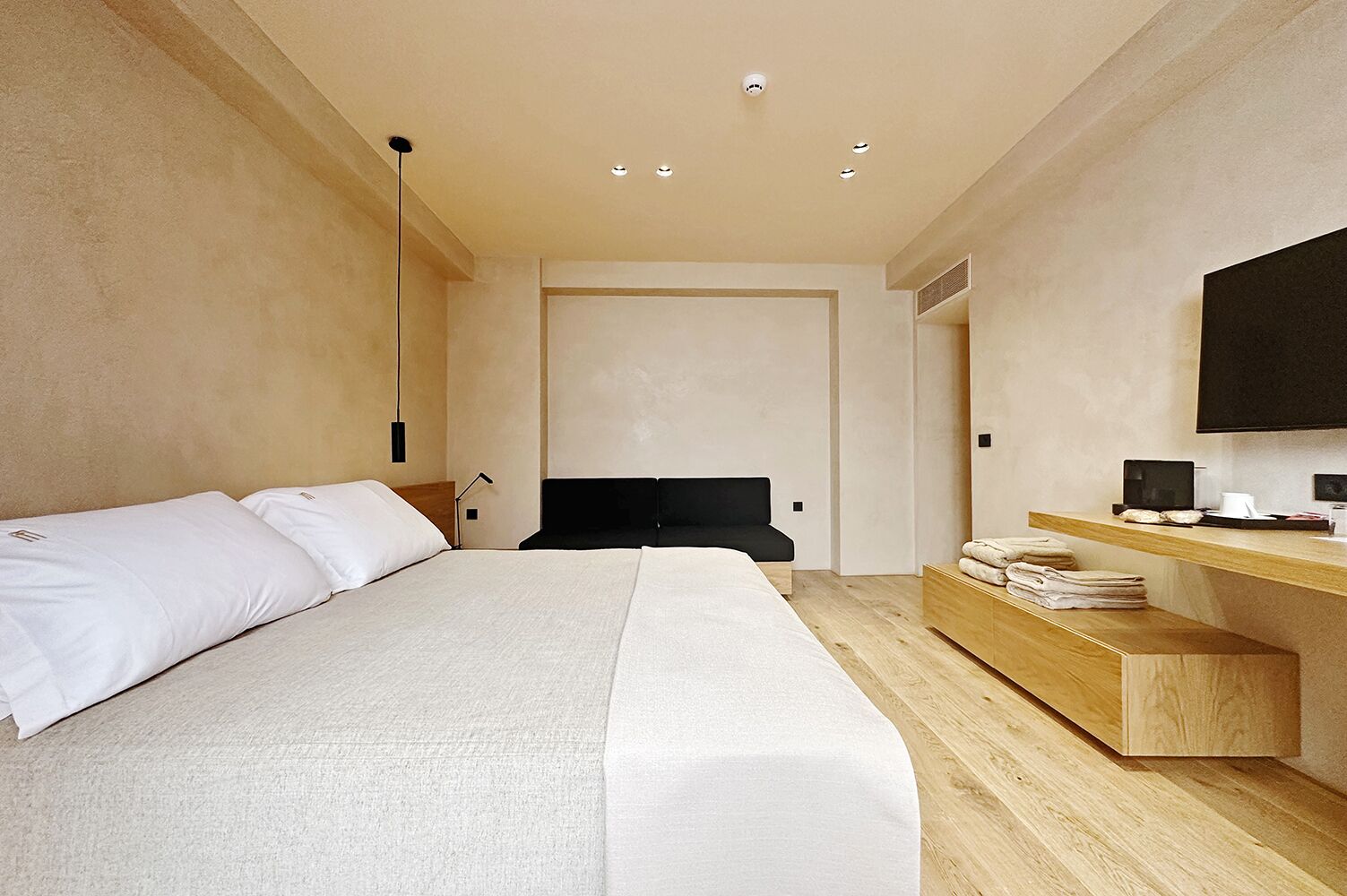 A modern hotel room with a large bed, a wooden bedside table, a flat-screen TV, and a built-in desk.
