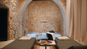 A spa room with a stone wall, a bathtub, a table with a bottle of champagne, glasses, and a wooden board with snacks. - Monastery Estate Venetian Harbor (Chania)