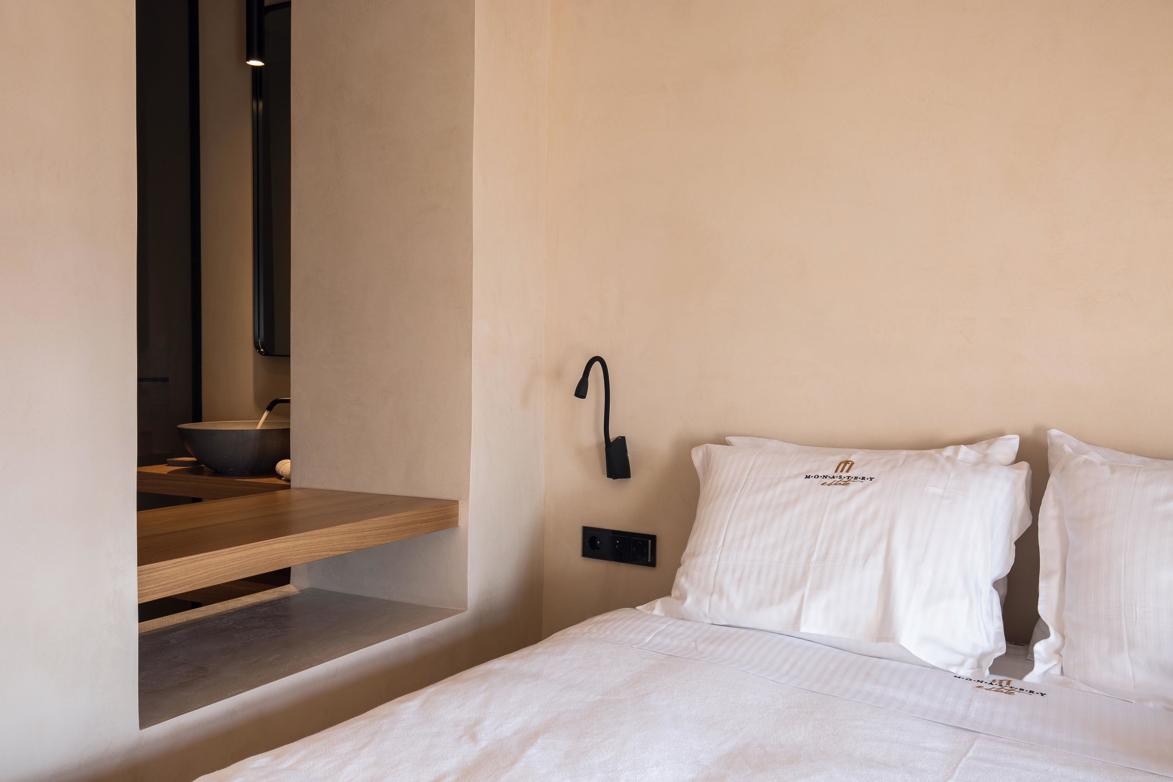 A minimalist hotel room with a wooden bedside table, a wall-mounted light, and a bed with white bedding.