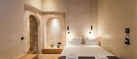 A modern hotel room with a wooden bench, a bed with white linens, a small plant, and a stone archway.