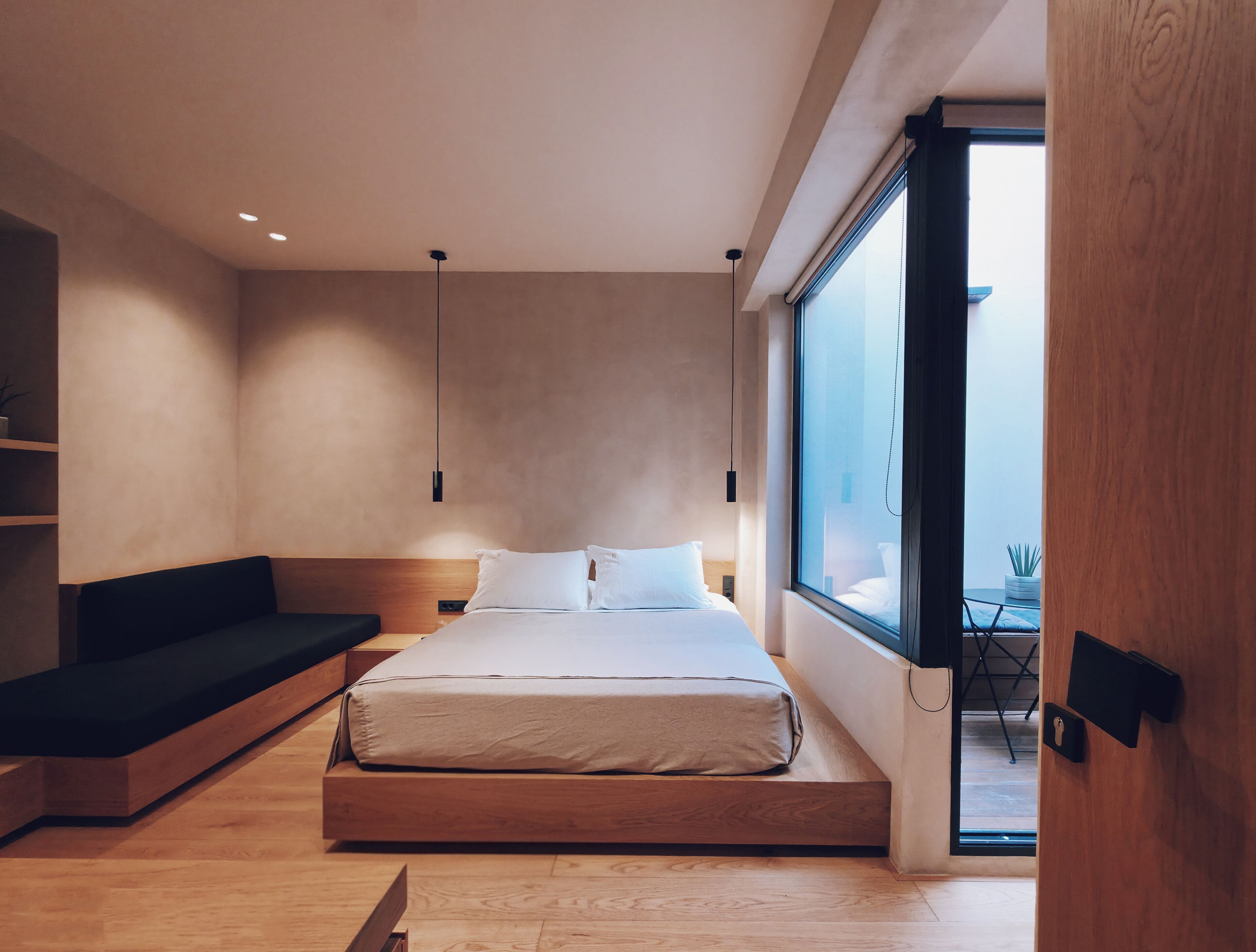 A modern bedroom with a minimalist design, featuring a bed with a wooden frame, a built-in bench, and a large window offering a view of the outdoors.