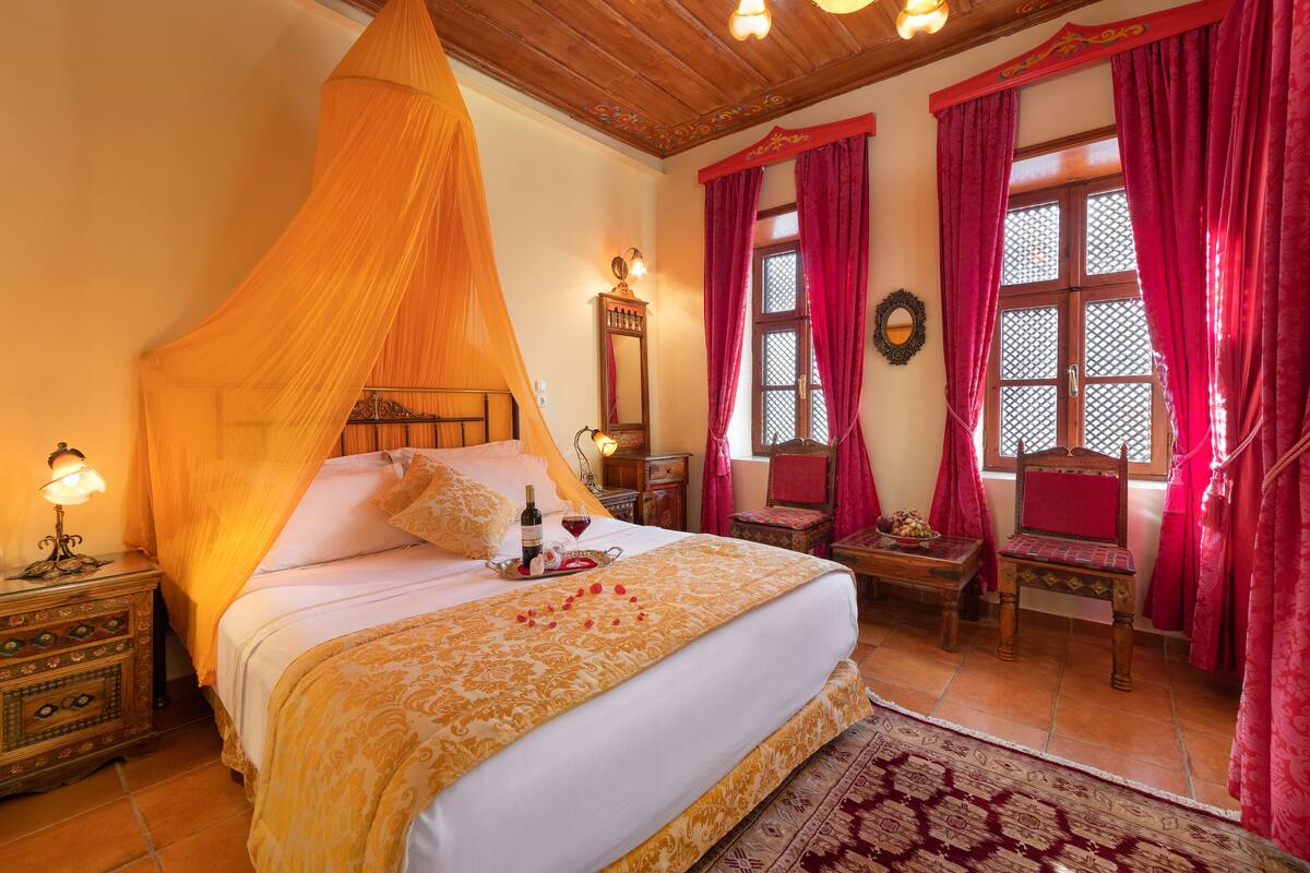 Un dormitorio con cama a dosel, una mesita de noche con lámpara, una silla de madera y una pequeña mesa con una bandeja de vino y flores.