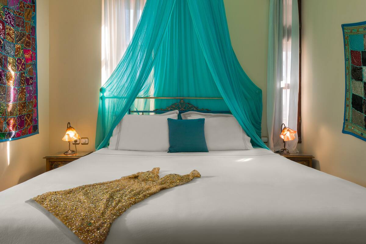 Una cama bien tendida con un dosel verde azulado, almohadas blancas y un adorno dorado.