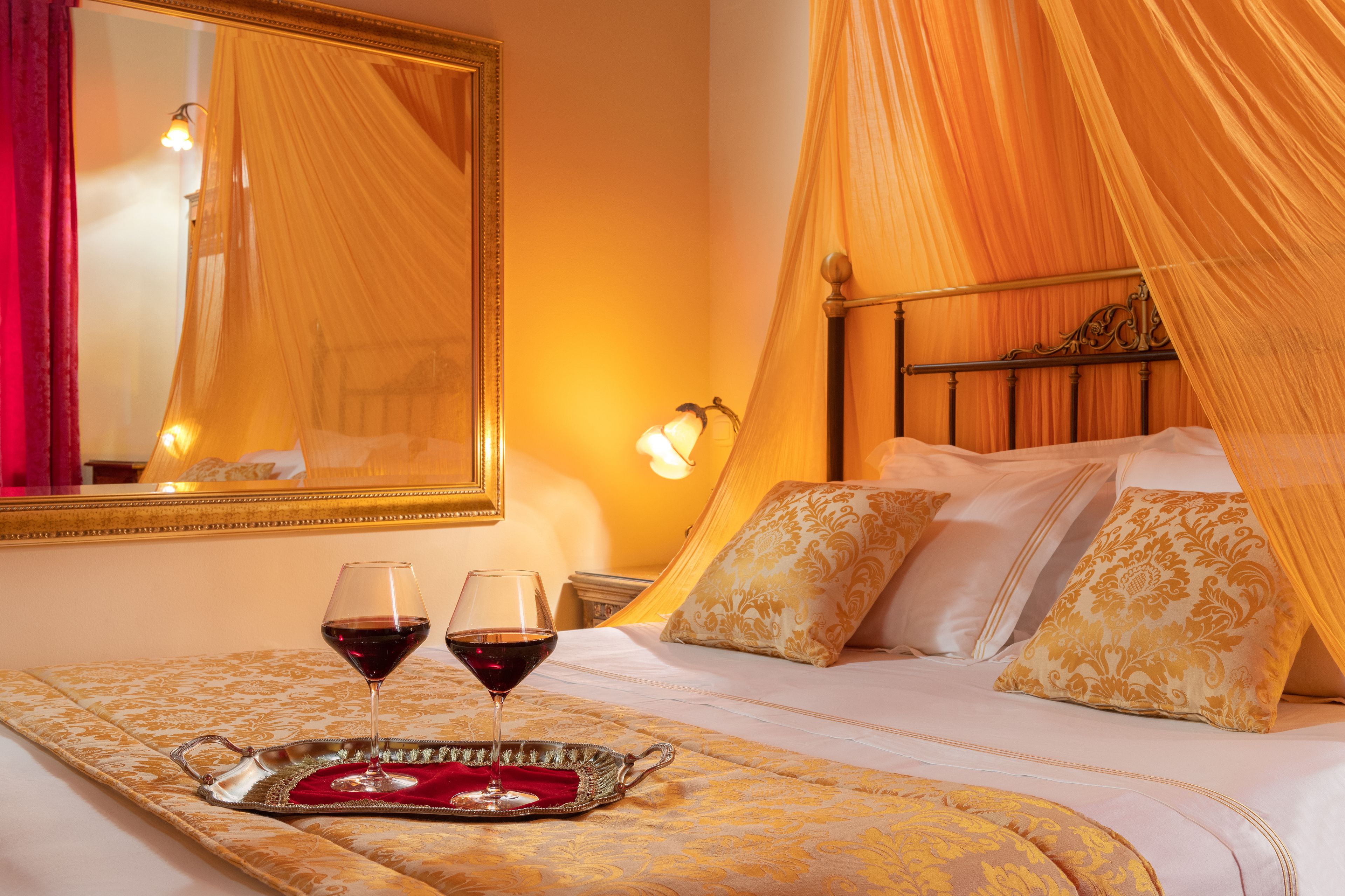 A hotel room with a bed, a canopy, a mirror, and two glasses of red wine on a tray.