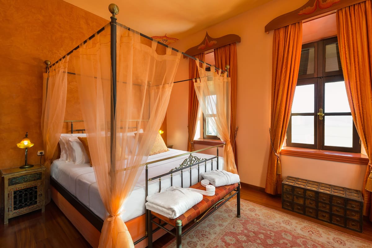 Una cama con dosel y cortinas, una mesita de noche con lámpara y una ventana con cortinas.
