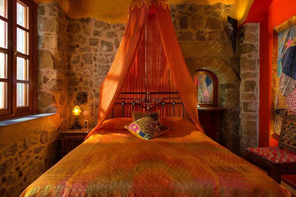 Un dormitorio con pared de piedra, cama con dosel, mesita de noche con lámpara y ventana con contraventanas de madera.