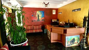 A reception area with a wooden desk, a sofa, a large potted plant, and a colorful painting on the wall. - Slightly Chilled Yellow House (Nallathanniya)