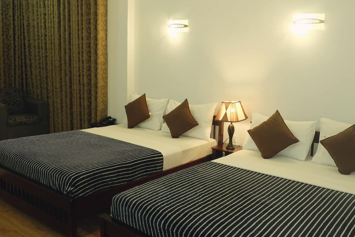 A hotel room with two beds, each with striped bedding and brown pillows. There is a nightstand with a lamp between the beds. The room has a curtain on one side and a wall-mounted light fixture above the beds.