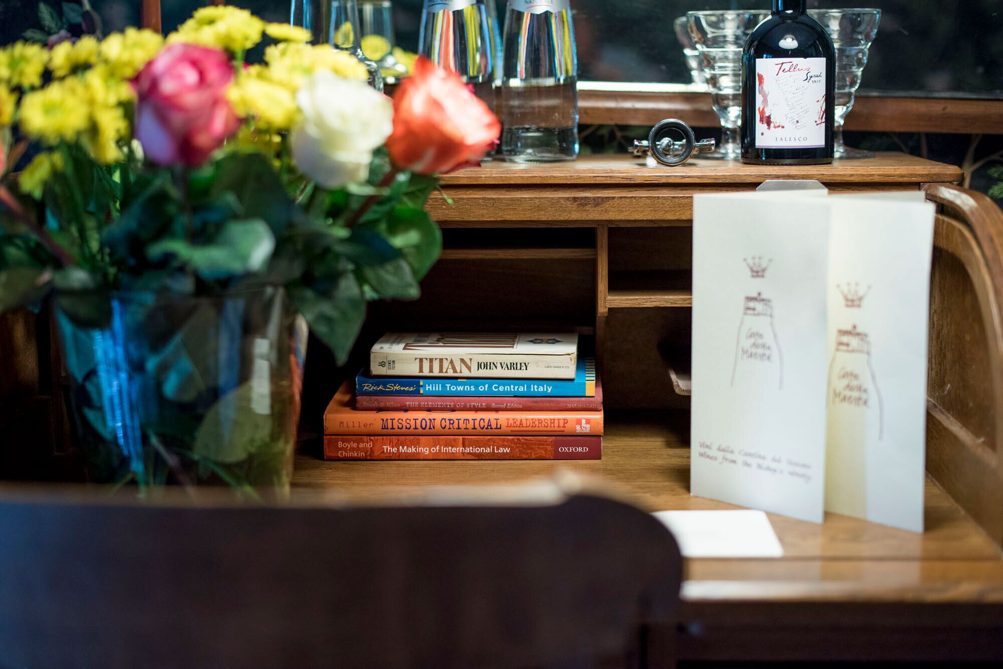 Uma escrivaninha de madeira com um vaso de flores, uma garrafa de vinho e vários livros.