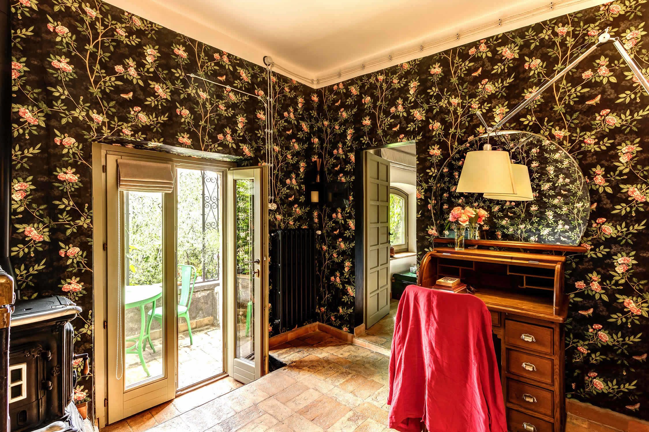 Une pièce avec un papier peint à motifs floraux, une commode en bois, une table recouverte d’une nappe rouge et une porte donnant sur un espace extérieur.
